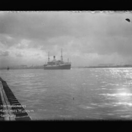 Dampfer im Hafen bei bewölktem Himmel, Pier links und Lichtreflexe auf dem Wasser; Logo des Maritimen Museums unten links.