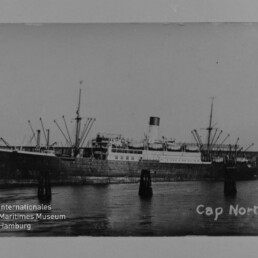 Schwarzweiß-Foto des Dampfschiffs Cap Norte mit Schornstein und Ladebäumen am Kai, mit Museumsstempel links unten.