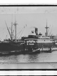 Historische Schwarzweißaufnahme eines großen Dampfschiffs im Hafen mit mehreren kleinen Booten daneben und Wasserzeichen 