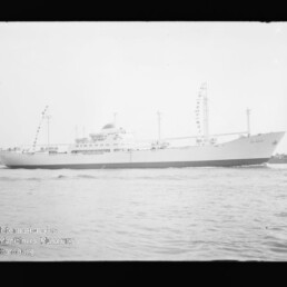 Weißes Passagier- bzw. Frachtschiff in Seitenansicht mit Masten und Signalflaggen, fahrend auf ruhigem Wasser.