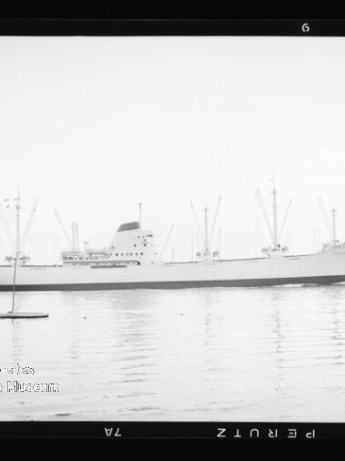 Großes Frachtschiff mit Ladekränen und Aufbauten auf ruhiger See, zwei kleine Segelboote davor.