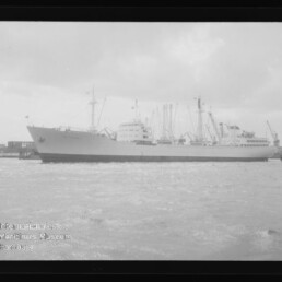 Großes Frachtschiff seitlich im Hafen vor Kai mit Ladekränen und einem Schlepper, schwarzweißfoto