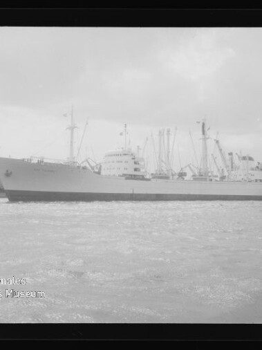 Großes Frachtschiff seitlich im Hafen vor Kai mit Ladekränen und einem Schlepper, schwarzweißfoto