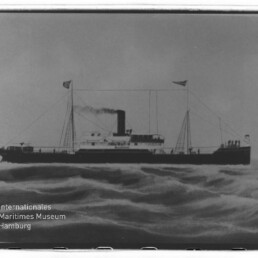Schwarzweißaufnahme eines Dampfschiffs mit hohem Schornstein und Flaggen, fahrend auf bewegter See, Museumswasserzeichen links.