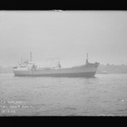 Schwarzweißaufnahme eines Frachtschiffs vor nebliger Hafenkulisse mit Kränen; Wasserzeichen Internationales Maritimes Museum Hamburg.