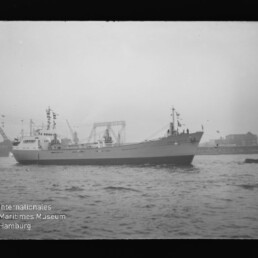 Schwarzweiß-Foto eines beflagten Frachtschiffs im Hafen vor Werftkränen, mit Wasser im Vordergrund und Museumslogo.