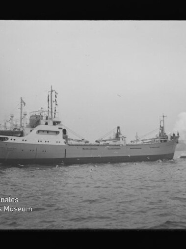 Frachtschiff mit vornliegenden Aufbauten und Flaggen, begleitet von kleinem Schlepper; sichtbares Museumslogo