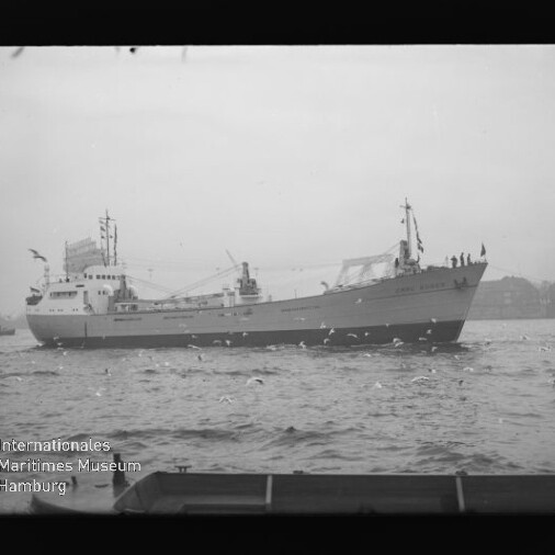 Frachtschiff auf ruhiger See mit fliegenden Möwen, Ufer im Hintergrund und sichtbarem Museumslogo, Schwarzweißaufnahme.