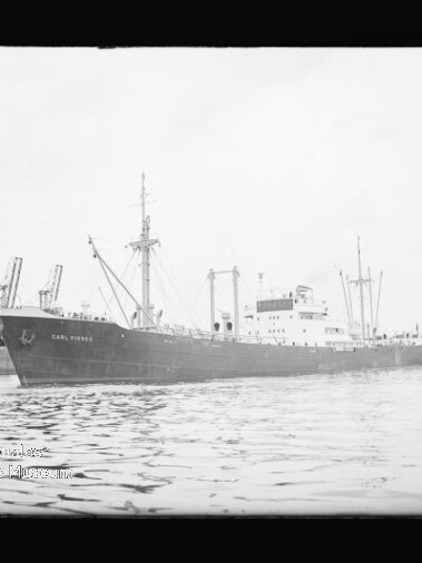 Großes Frachtschiff vor Kaianlagen mit Hafenkränen, spiegelndes ruhiges Wasser.