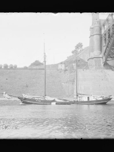 Zweimastiges Segelschiff liegt auf ruhigem Fluss vor massivem Brückenpfeiler und Hügel mit einer Baumreihe im Hintergrund.