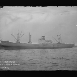 Schwarzweißfoto eines Frachtschiffs mit Ladekränen auf stillem Meer, Museumslogo unten links.