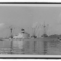 Frachtschiff mit Kränen und Masten auf ruhigem Wasser, Schwarzweißfoto mit Wasserzeichen des Maritimen Museums Hamburg.