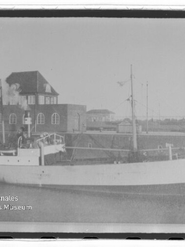 Weißes Frachtschiff am Kai vor Backsteinhaus mit Rundfenstern; unten links Logo Internationales Maritimes Museum Hamburg.