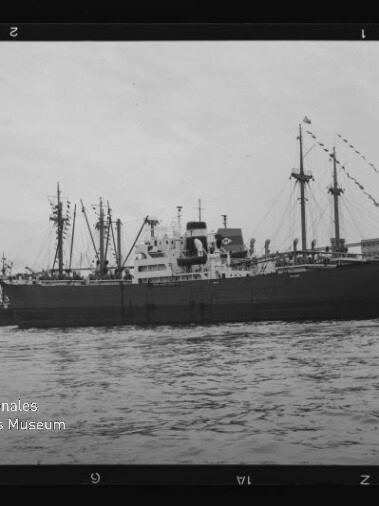 Schwarzweiß-Foto eines großen Frachtschiffs im Hafen, Wasser im Vordergrund und das Logo „Internationales Maritimes Museum Hamburg“.