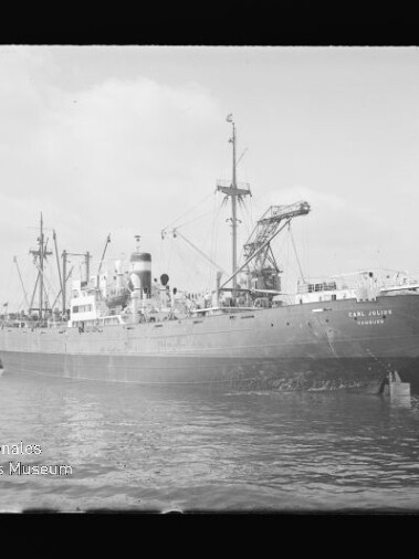 Großes Frachtschiff mit Deckskranen am Kai eines Industriehafens, ruhiges Wasser im Vordergrund.