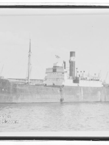 Schwarzweiß-Seitenansicht eines dampfgetriebenen Frachtschiffs mit Schornstein und zwei Masten im Hafen.
