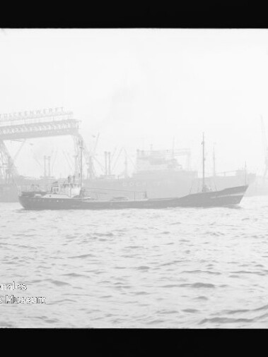 Frachtschiff fährt in nebligem Hafen vor einer Werft mit großem Kran und mehreren Schiffen im Hintergrund.