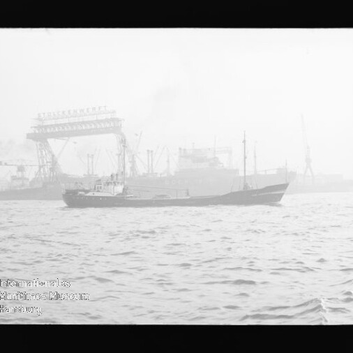 Frachtschiff fährt in nebligem Hafen vor einer Werft mit großem Kran und mehreren Schiffen im Hintergrund.