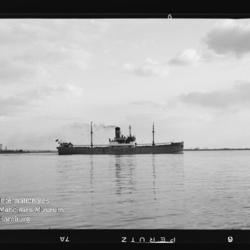 Schwarzweißfoto eines Frachtdampfers mit Schornstein und Masten auf ruhigem Wasser, mit Spiegelung und Museums-Wasserzeichen.