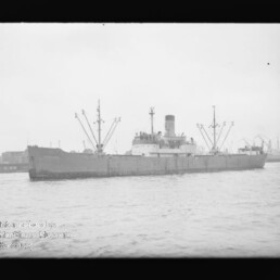Frachtschiff mit hohem Schornstein und mehreren Ladebäumen auf dem Wasser, dahinter Hafenpier und Kräne.