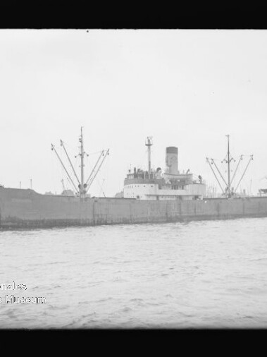 Frachtschiff mit hohem Schornstein und mehreren Ladebäumen auf dem Wasser, dahinter Hafenpier und Kräne.