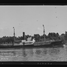 Schwarzweißfoto eines kleinen Dampfers mit Schornstein auf einem Fluss vor bewaldetem Ufer und Häusern.