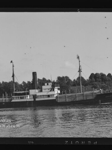 Schwarzweißfoto eines kleinen Dampfers mit Schornstein auf einem Fluss vor bewaldetem Ufer und Häusern.