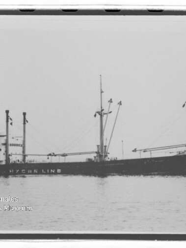 Schwarzweiß-Seitenansicht eines Frachtschiffs mit Ladebäumen und Masten auf ruhiger See; Wasserzeichen des Maritimen Museums Hamburg