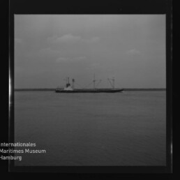 Frachtschiff mit zwei Masten und Schornstein auf ruhiger See vor flachem Horizont; Museumslogo und Schriftzug unten links.