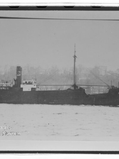 Seitliches Schwarz-Weiß-Bild eines Frachtschiffs auf ruhiger See mit Stadtsilhouette im Hintergrund und Museumswasserzeichen.