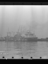 Mehrere angelegte Frachtschiffe und Kräne in nebligem Hafen, ruhige Wasserfläche; Museumssignet unten links.