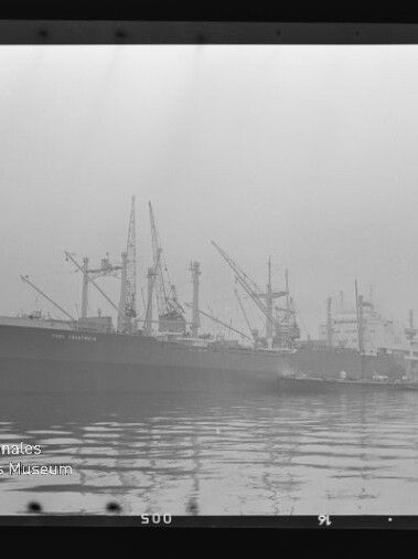 Großes Frachtschiff am Kai zwischen Hafenkränen, Spiegelung und ruhigem Wasser in einer schwarz-weißen Aufnahme.