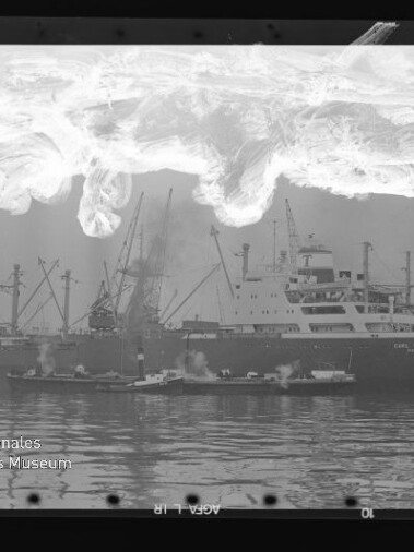 Frachtschiff mit Kränen am Kai, Schlepper im Vordergrund, Spiegelung im Wasser und helle Filmschäden oben; Museumswasserzeichen.