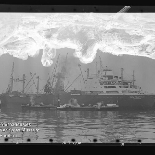 Frachtschiff mit Kränen am Kai, Schlepper im Vordergrund, Spiegelung im Wasser und helle Filmschäden oben; Museumswasserzeichen.