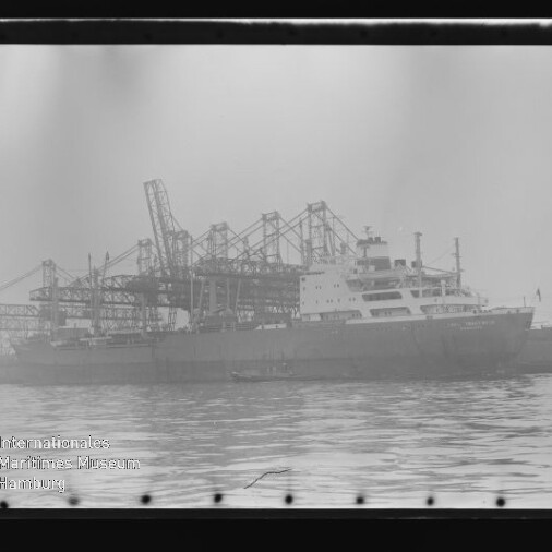 Großes Frachtschiff am Industriehafen mit mehreren Ladekränen im Hintergrund und ruhiger Wasserfläche im Vordergrund.