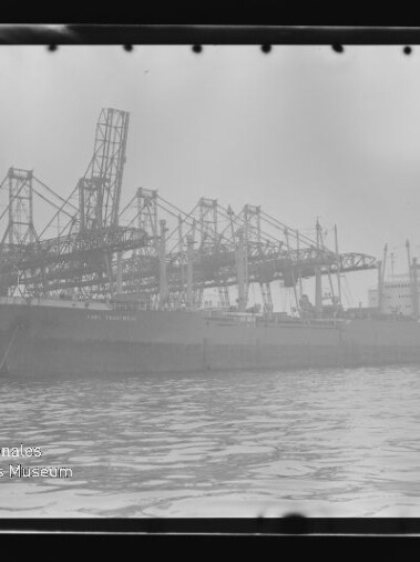 Frachtschiff am Kai unter hohen Verladekränen, ruhiges Wasser im Vordergrund und Wasserzeichen Internationales Maritimes Museum Hamburg.