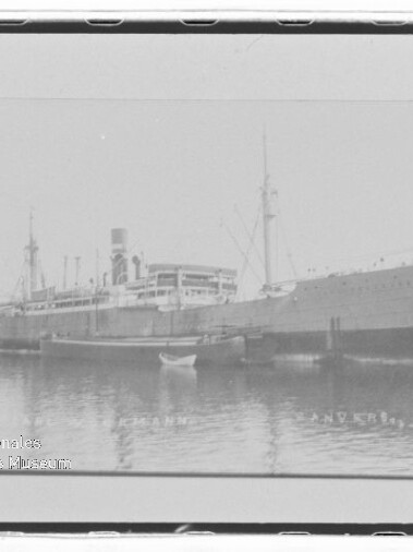 Schwarzweißaufnahme eines großen historischen Dampfschiffs am Kai, mit Spiegelung im Wasser und kleinem Ruderboot im Vordergrund.
