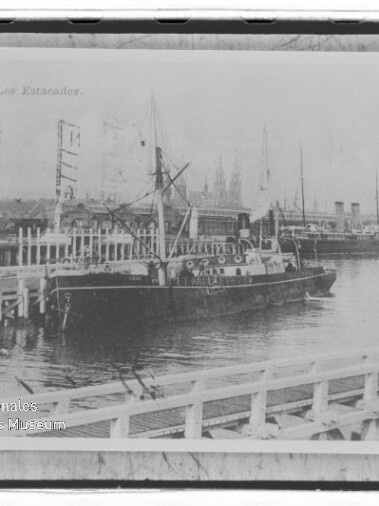 Schwarzweißaufnahme eines Dampfschiffs am hölzernen Kai in Ostende; oben links Beschriftung OSTENDE — Les Estacades, Masten und Geländer sichtbar.