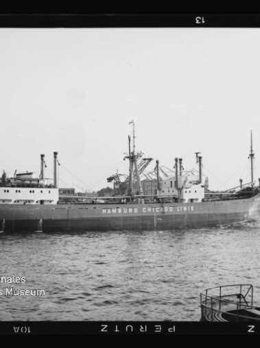 Schwarzweißfoto eines Frachtschiffs mit der Aufschrift HAMBURG CHICAGO LINE im Hafen, Kräne am Ufer.