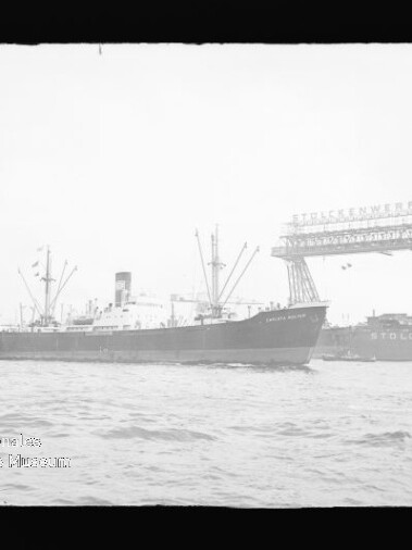 Frachtschiff passiert eine Werft mit großer Auslegerbrücke im Hafen, Wasser im Vordergrund.
