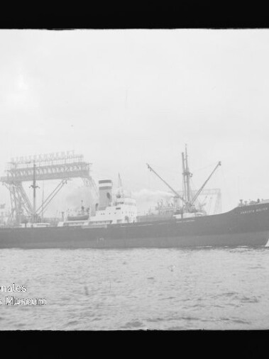 Frachtschiff im Hafen vor großen Krananlagen; links unten Logo mit Schrift Internationales Maritimes Museum Hamburg.