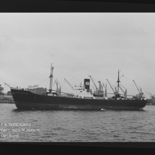 Schwarzweiß-Seitenansicht eines Frachtschiffs im Hafen vor zahlreichen Ladekränen, Museumslogo unten links.