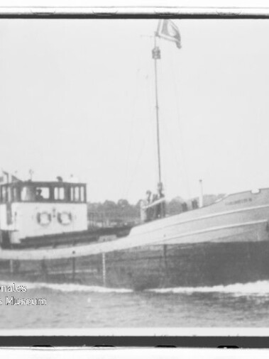 Kleiner Frachter mit Bug rechts, Steuerhaus und Mast, vor Ufer; Wz. Maritimes Museum Hamburg