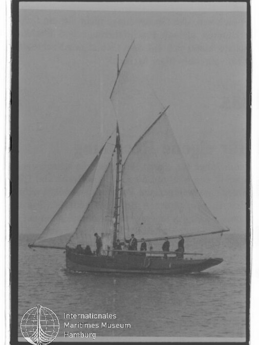 Segelboot mit zwei Segeln und mehreren Personen an Bord auf ruhiger See; Schwarzweißfoto mit Museumswasserzeichen