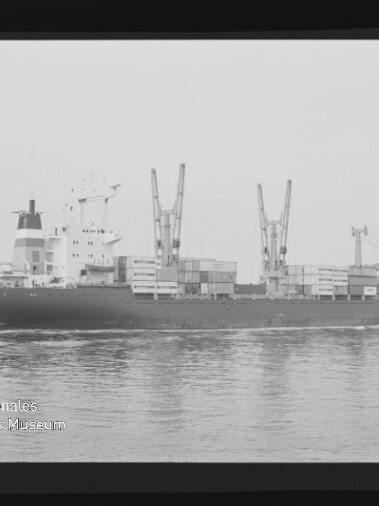 Schwarzweißfoto eines Frachtschiffs mit Ladekränen und Containern auf ruhiger See, Filmrand sichtbar.