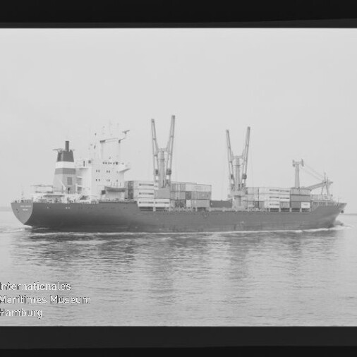 Schwarzweißfoto eines Frachtschiffs mit Ladekränen und Containern auf ruhiger See, Filmrand sichtbar.