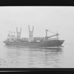 Containerschiff mit Containern und Ladekränen fährt auf ruhiger See; schwarzweiß-Foto mit Museumswasserzeichen.