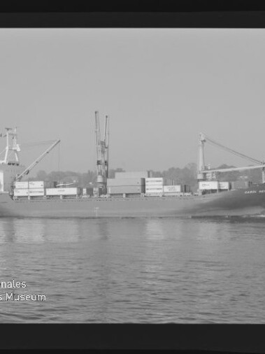 Schwarzweißaufnahme eines Containerschiffs mit Deckkränen und Containern, fahrend auf ruhigem Wasser, Wasserzeichen links.