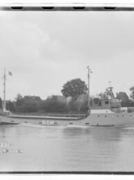 Schwarzweiß-Seitenansicht eines Frachtschiffs mit Kennzeichnung STM, fahrend auf ruhigem Fluss vor bewaldetem Ufer und Häusern.