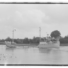 Schwarzweißaufnahme eines kleinen Frachtschiffs mit Schriftzug STN auf ruhigem Fluss, Bäume und Häuser im Hintergrund; Wasserzeichen des Internationalen Maritimen Museums Hamburg.